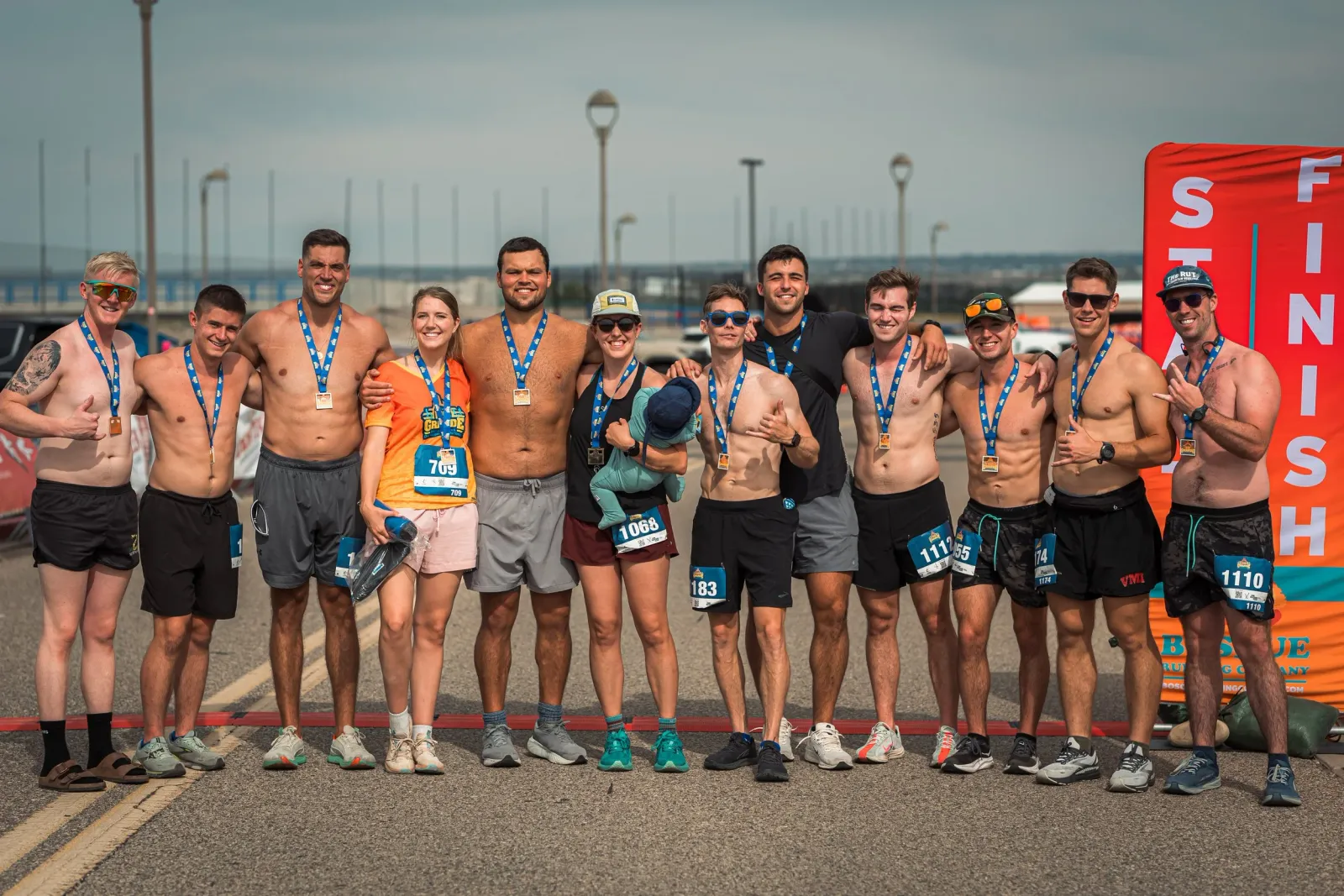 A group of runners, some shirtless and wearing medals, pose for a photo in front of a finish line banner.