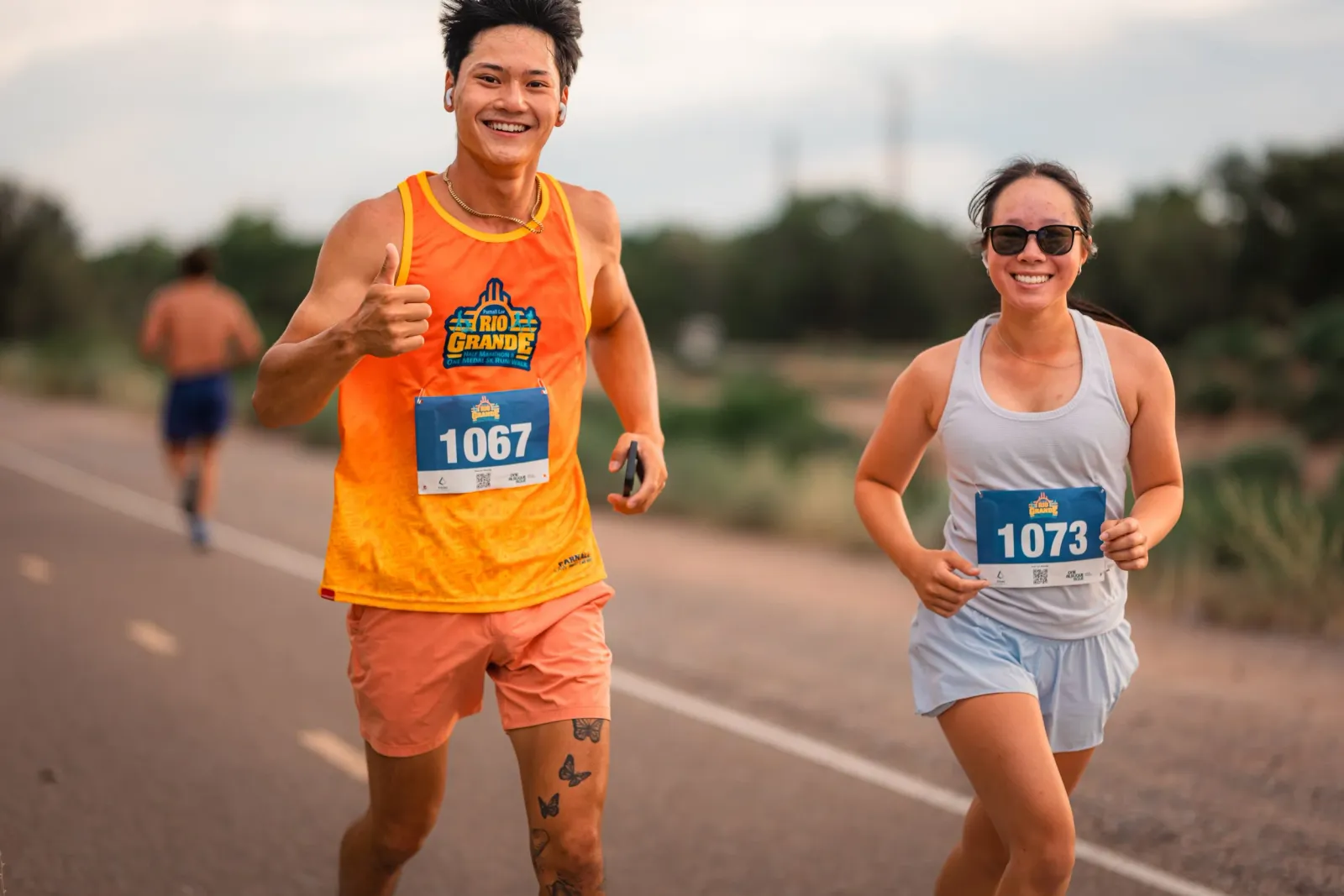 A male runner gives a thumbs up while running, and a female runner smiles as they both participate in the Rio Grande Half Marathon.