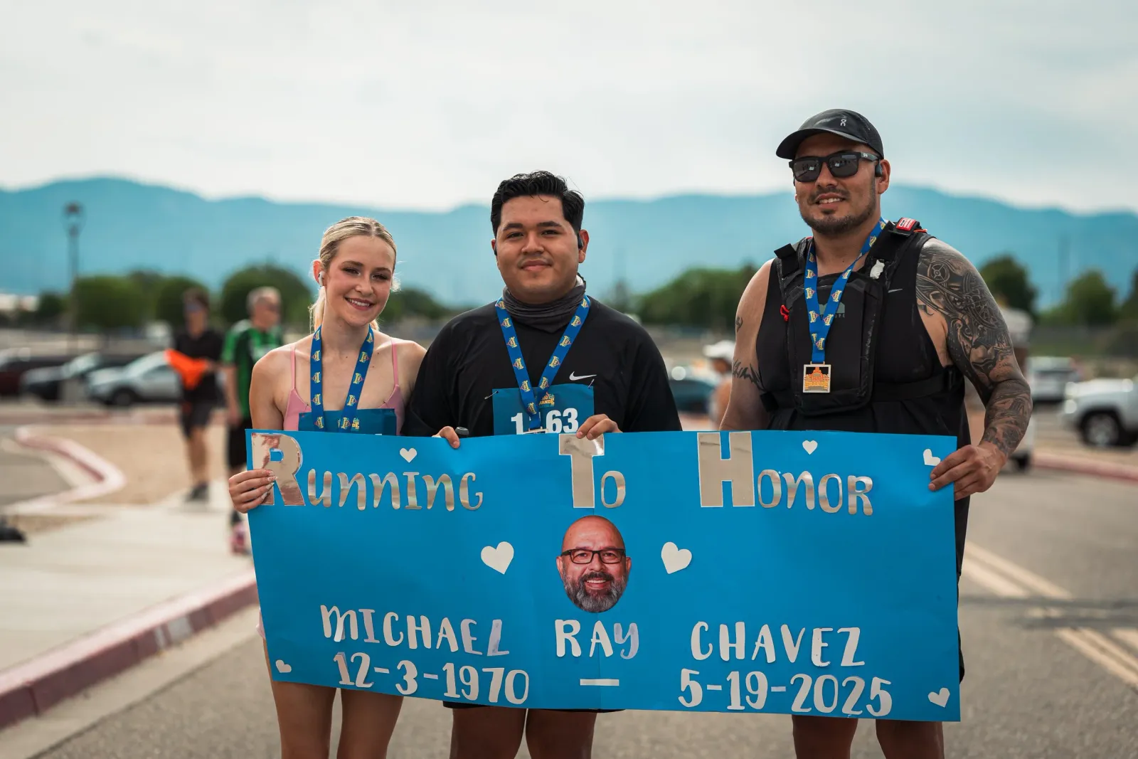 Three people smiling and holding a blue banner that reads 'Running To HONOR MICHAEL RAY CHAVEZ 12-3-1970 - 5-19-2025', with a photo of Michael Ray Chavez on the banner.