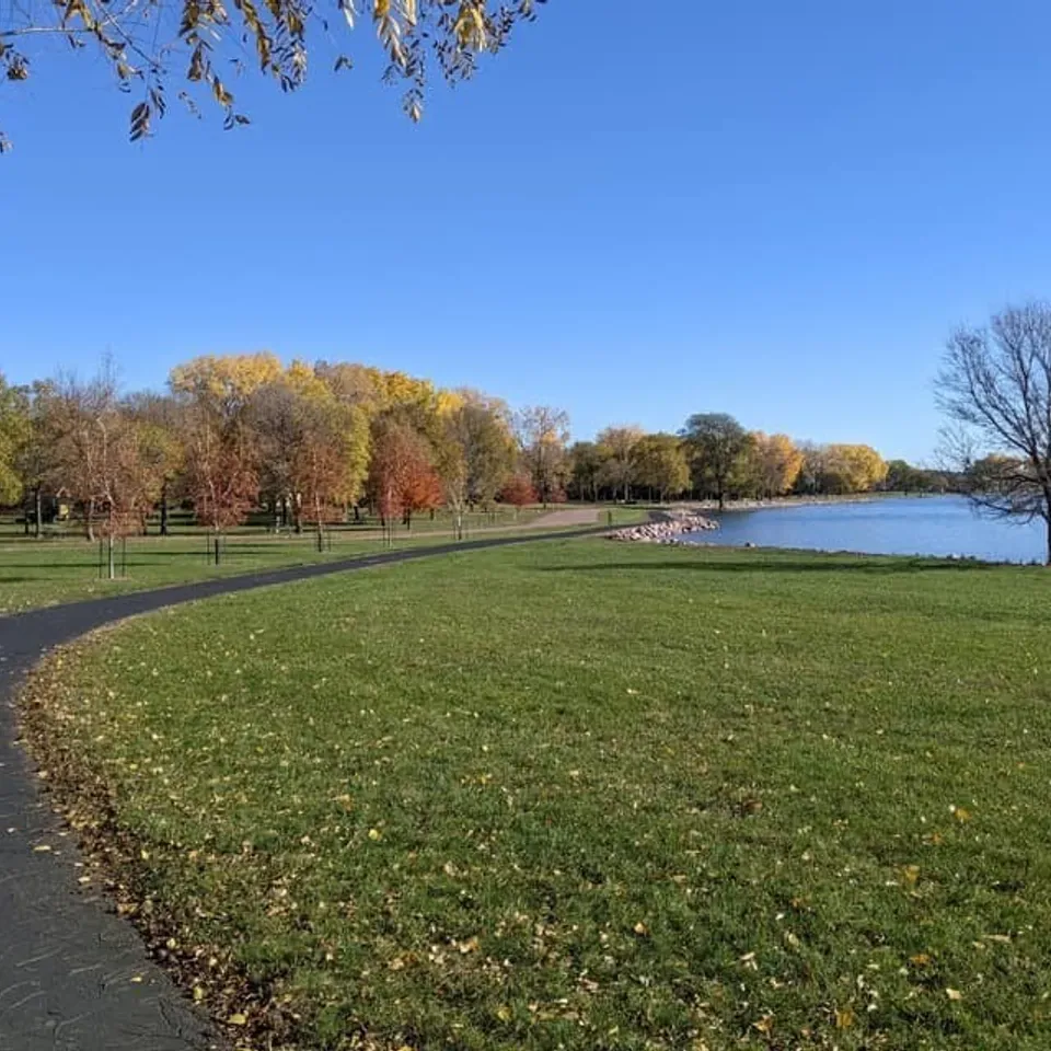 A paved path winds through a grassy park next to a calm body of water, with trees displaying autumn foliage under a clear blue sky.