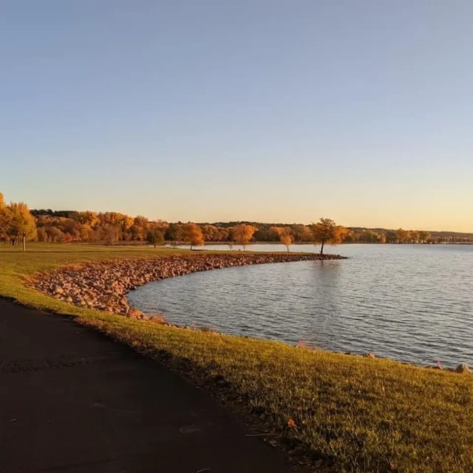 A paved path curves along the edge of a large body of water, with a rocky shoreline and trees displaying autumn colors in the background under a clear sky.