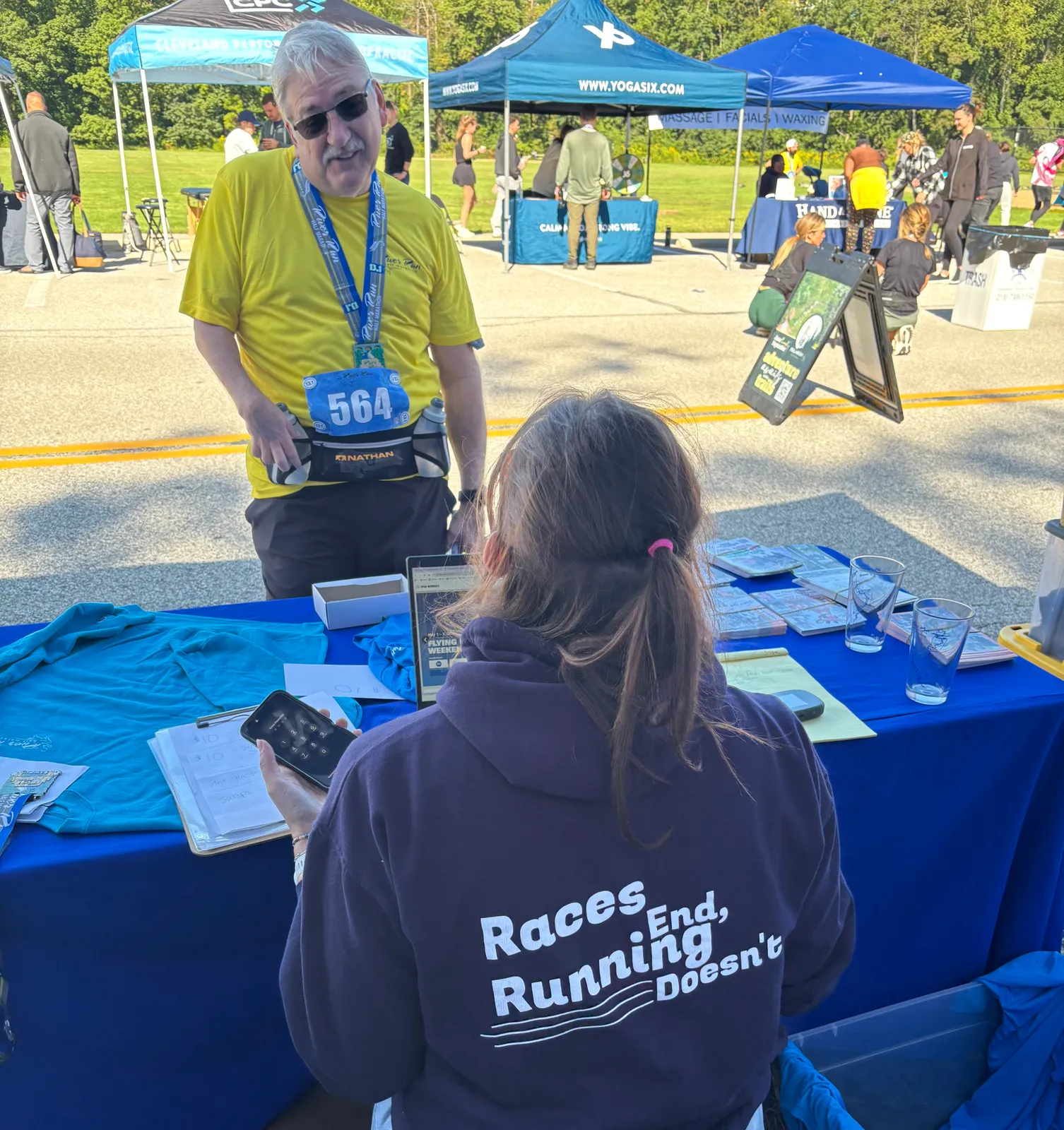 A runner wearing a yellow shirt and a medal talks to someone behind a table at a race event.
