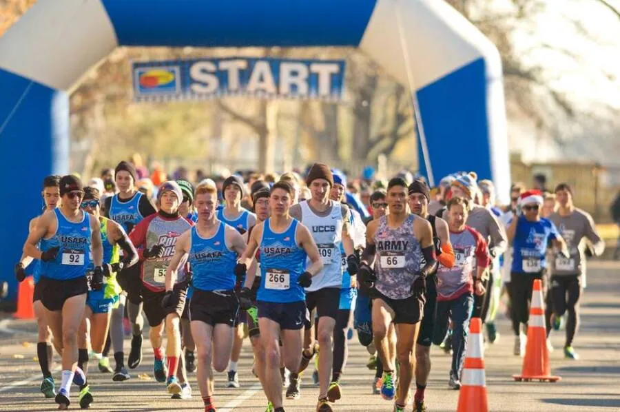 Runners in athletic wear are starting a race, with a blue and white archway labeled 'START' overhead. Many runners are wearing blue singlets with 'USAFA' printed on them, and some have bib numbers visible.