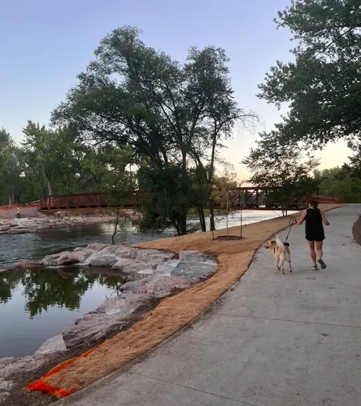 A woman runs with her dog on a paved path alongside a river, with a bridge in the distance.