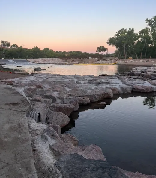 Scenic view of a rocky waterfront at sunset, with a dam or weir in the distance.