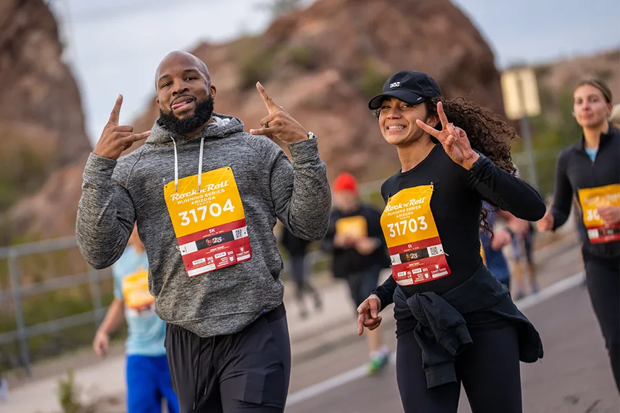Two runners, a man and a woman, smiling and making celebratory gestures while running in a race, with rocky hills in the background.