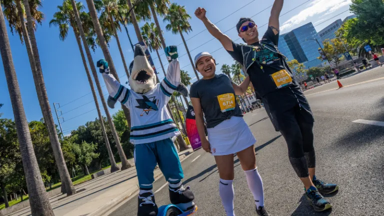 A mascot and two runners celebrating with their arms in the air on a race course lined with palm trees.