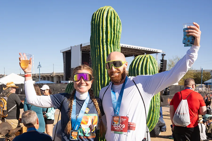 A smiling woman and man, both wearing race medals and holding drinks, pose in front of large cactus decorations at the Rock 'n' Roll Arizona Marathon.