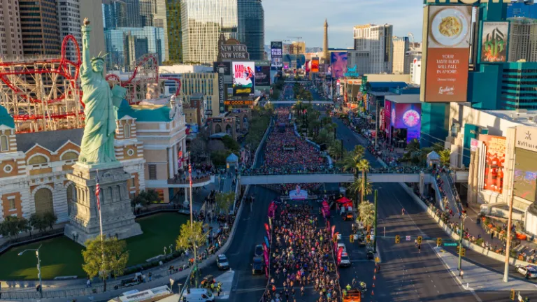 A large crowd of runners participating in a marathon on the Las Vegas Strip, with iconic hotels and the Statue of Liberty replica in the background.