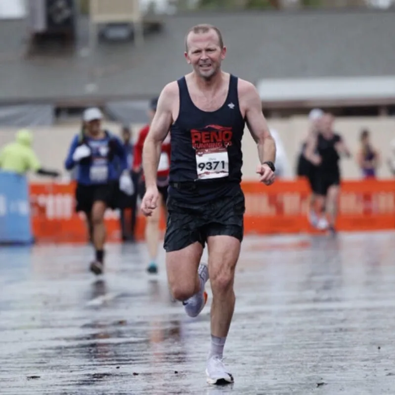 A male runner wearing a black tank top with 'RENO RUNNING' and bib number 9371, running on a wet road during a race.