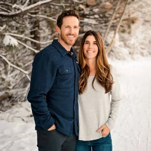A smiling couple standing in a snowy, wooded area.