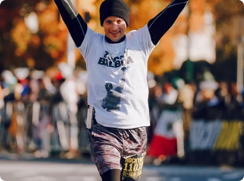 A runner with arms raised in celebration, wearing a Rocky Balboa t-shirt, crosses the finish line of a race.