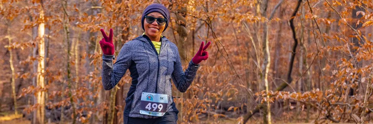 A female runner wearing a beanie, sunglasses, and a grey jacket smiles and holds up two peace signs while running in a forest with autumn foliage.