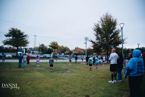 A group of runners and spectators gathered on a grassy area with trees and parked cars in the background.
