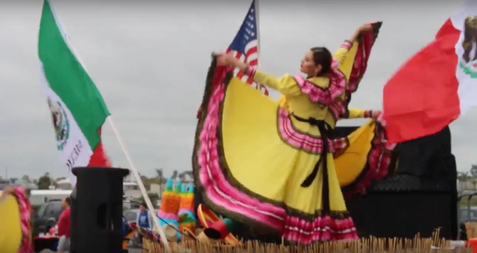 A woman in a yellow and pink traditional dress dances with flags, including the Mexican and American flags, at an outdoor event.