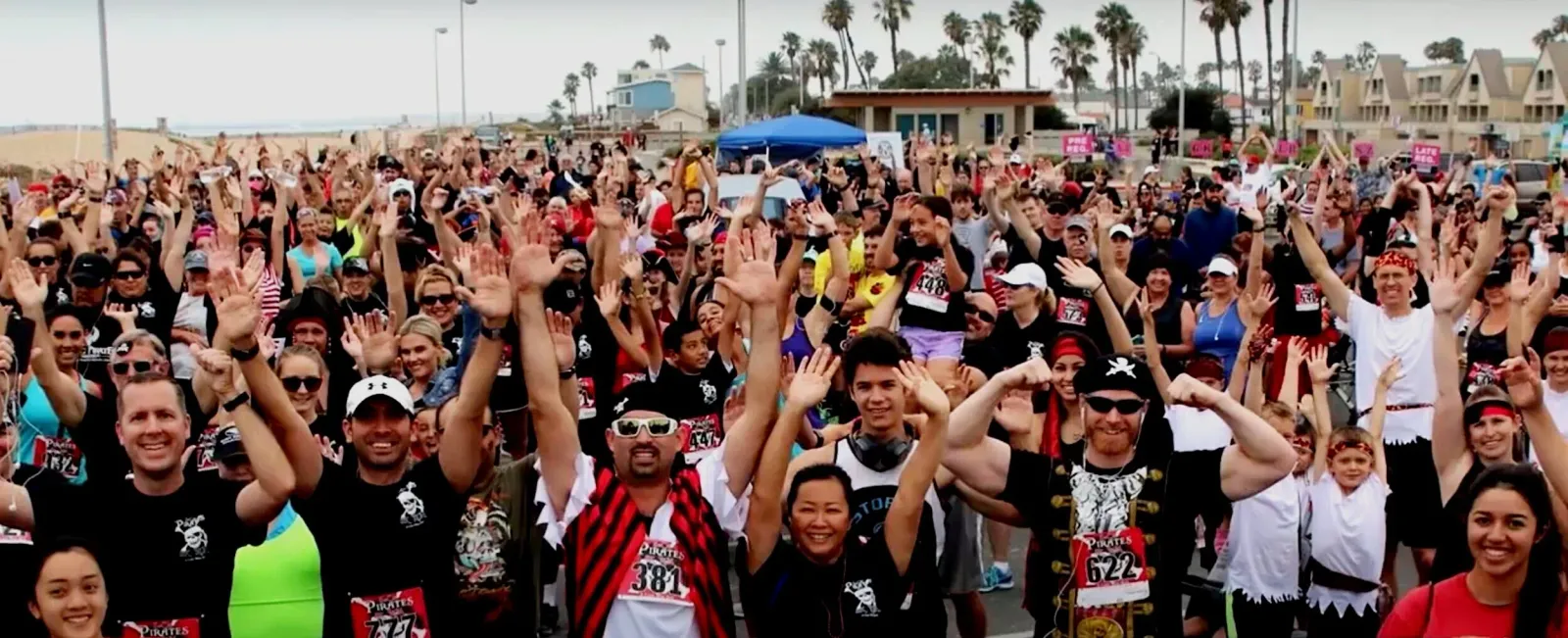 A large, diverse crowd of runners with their hands raised in excitement at a race event.