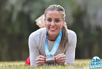 A smiling woman lies on the grass, holding up her race medal from Run Jekyll.