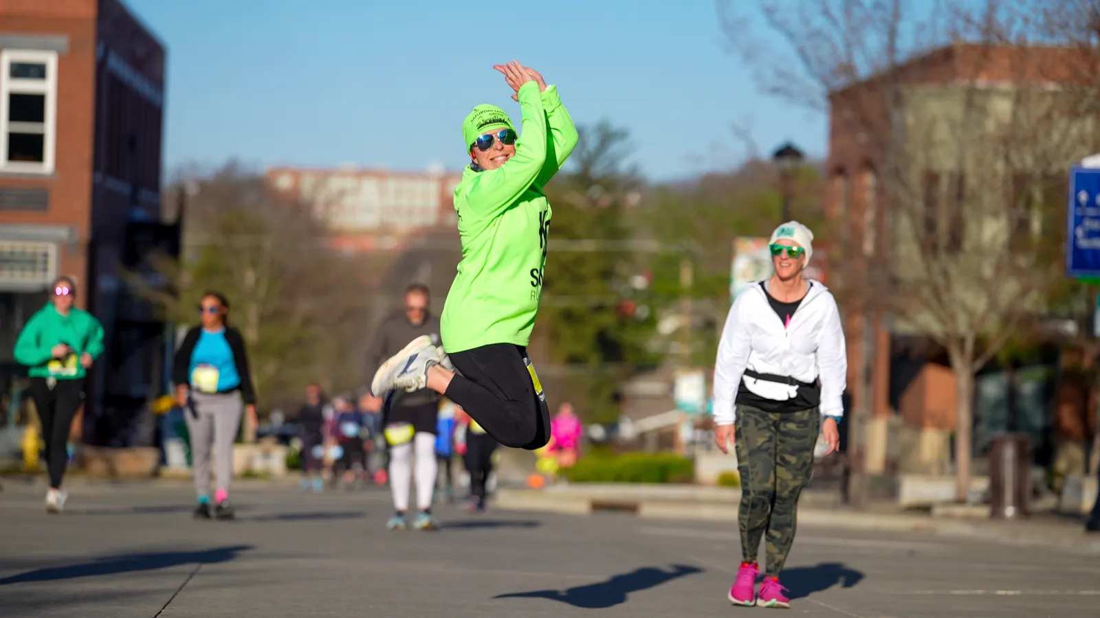 A runner in a bright green shirt and hat jumps in the air with a smile during a race, with other runners and buildings in the background.