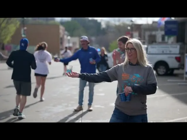 A volunteer hands a cup to a runner during a race, with other runners and spectators in the background.