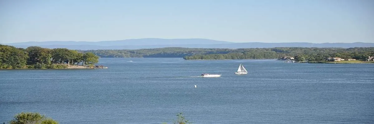 Scenic view of a wide blue lake with sailboats and a motorboat, bordered by tree-covered hills and distant mountains under a clear blue sky.