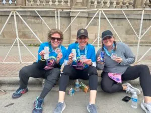 Three women sitting on the ground, smiling and holding up their race medals.