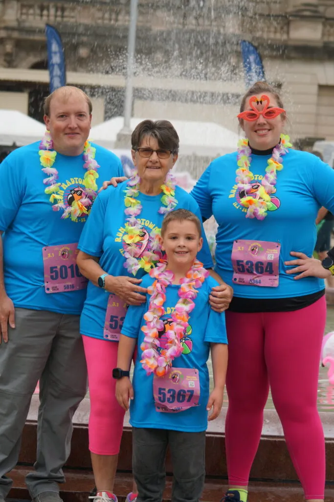 A family of four, including two adults, a grandmother, and a young boy, pose for a photo wearing blue race t-shirts, colorful leis, and pink leggings. The boy and one adult wear fun flamingo-shaped glasses. Bib numbers are visible on their shirts.