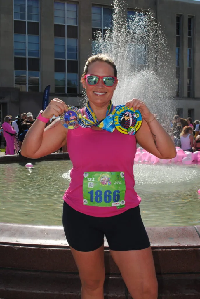 A woman in a pink tank top and black shorts smiles and holds up two race medals in front of a fountain.