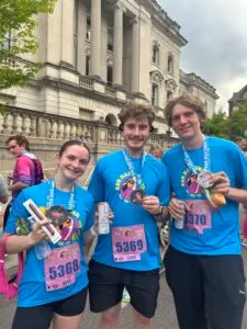 Three smiling runners, two men and one woman, wearing blue race t-shirts and medals, pose for a photo after completing a race.