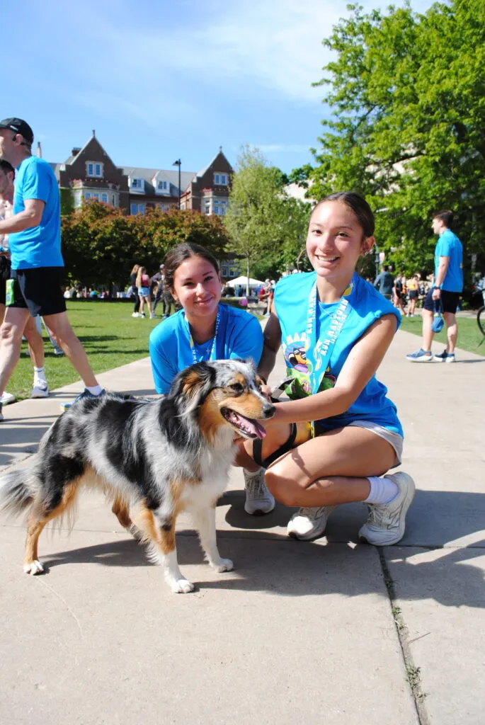 Two smiling young women wearing race medals and blue t-shirts pose with a dog on a sunny day, with other participants and buildings in the background.