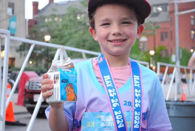 A young boy smiles at the camera, wearing a race medal and holding a carton of chocolate milk.