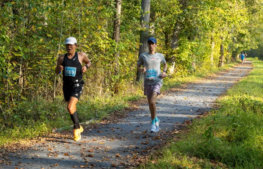 Two runners on a paved path through a forest, with trees and foliage on either side.