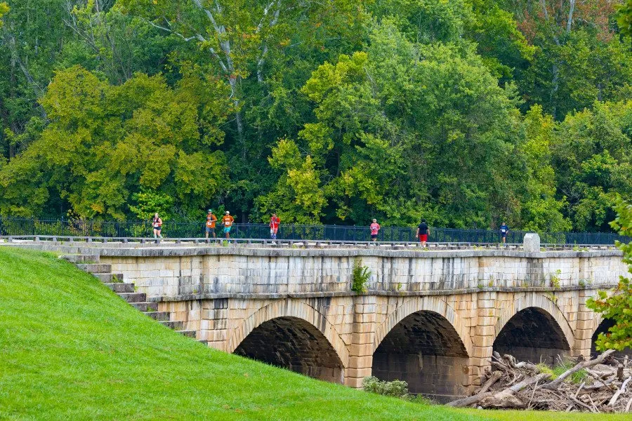 Runners crossing a stone bridge with lush green trees in the background.