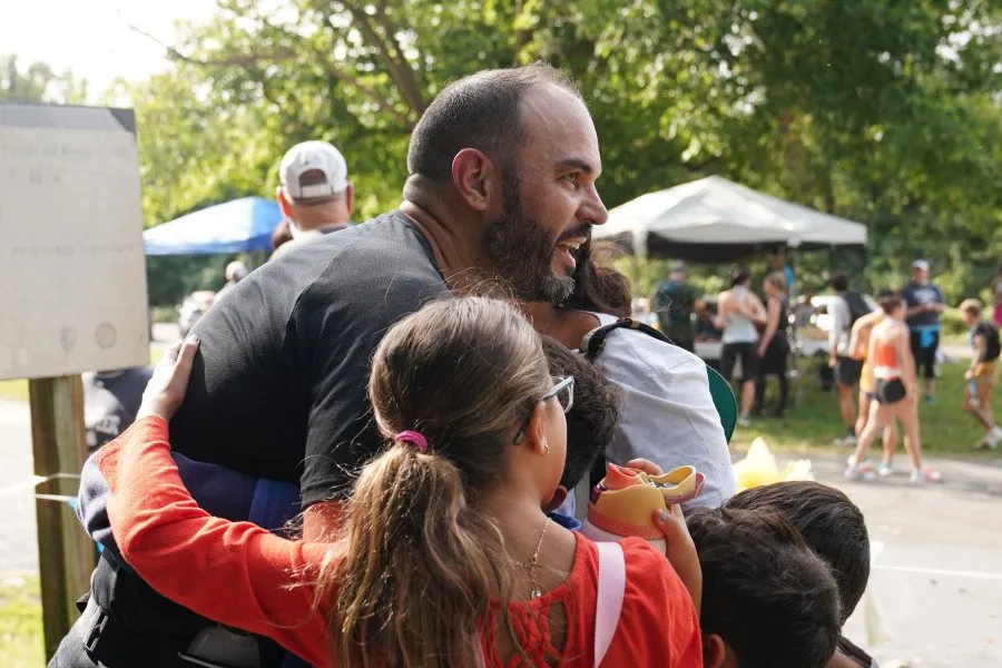 A runner with a beard is joyfully embracing children after a race, with other participants and tents visible in the background.