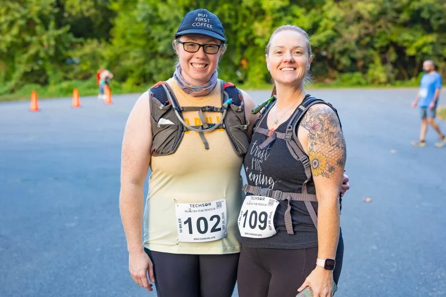 Two women wearing running bibs and hydration vests smile at the camera, with other runners and orange cones visible in the background.