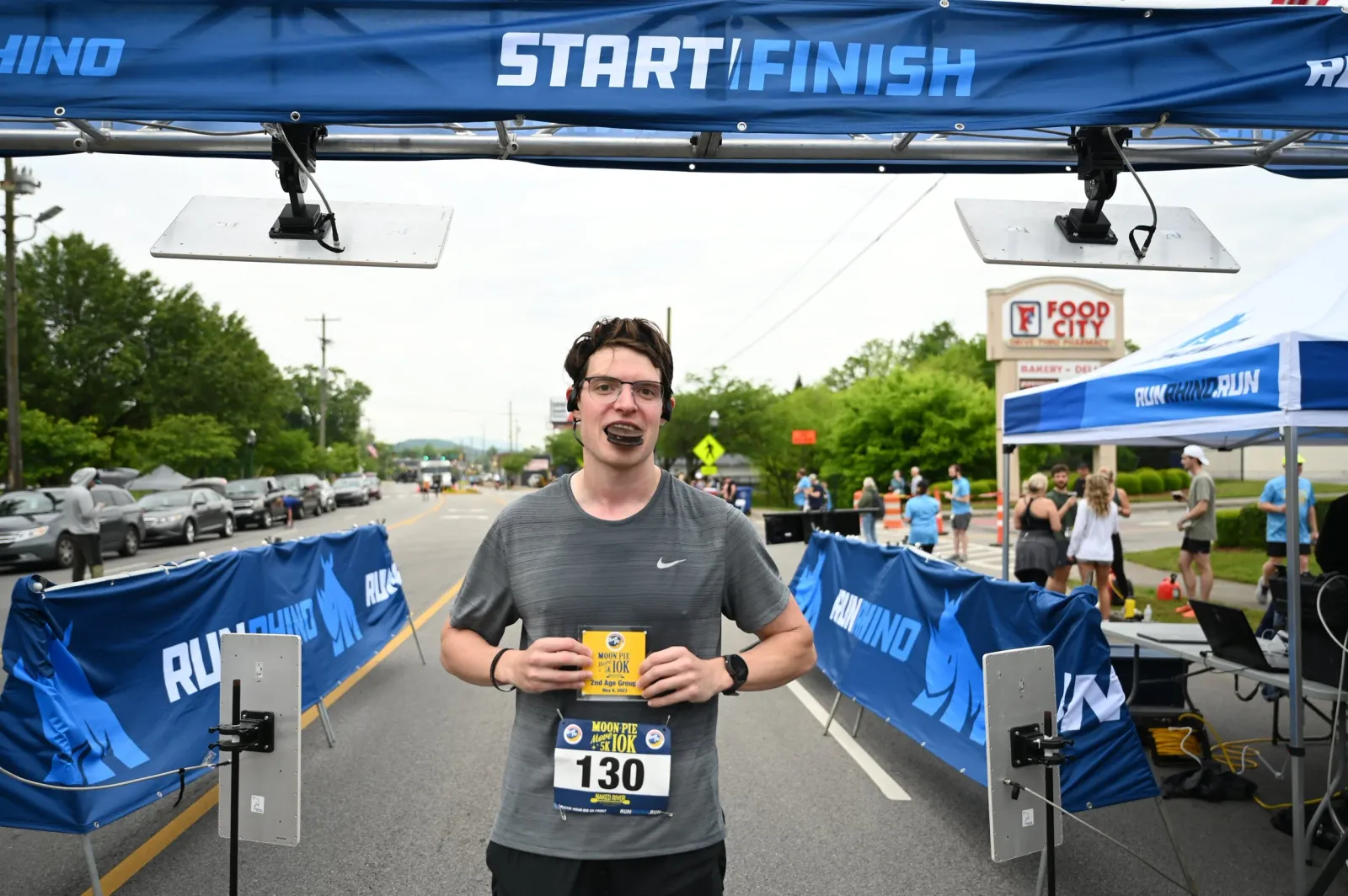 A runner smiles at the camera, holding a race bib and a small award, with a 'START/FINISH' banner overhead and other runners in the background.