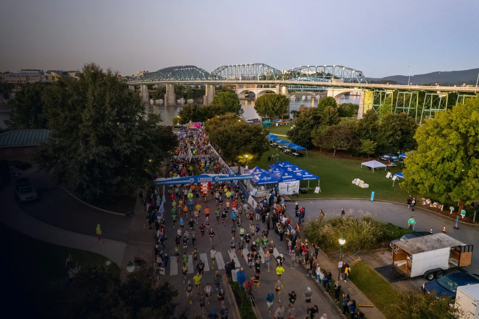 Runners crossing the start line of the 7 Bridges Marathon with a large bridge and river in the background.