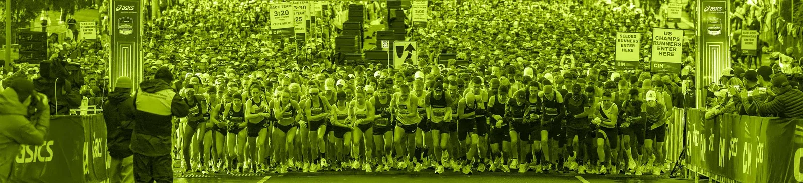 A large crowd of runners at the start line of a race, with pace team signs visible in the background.
