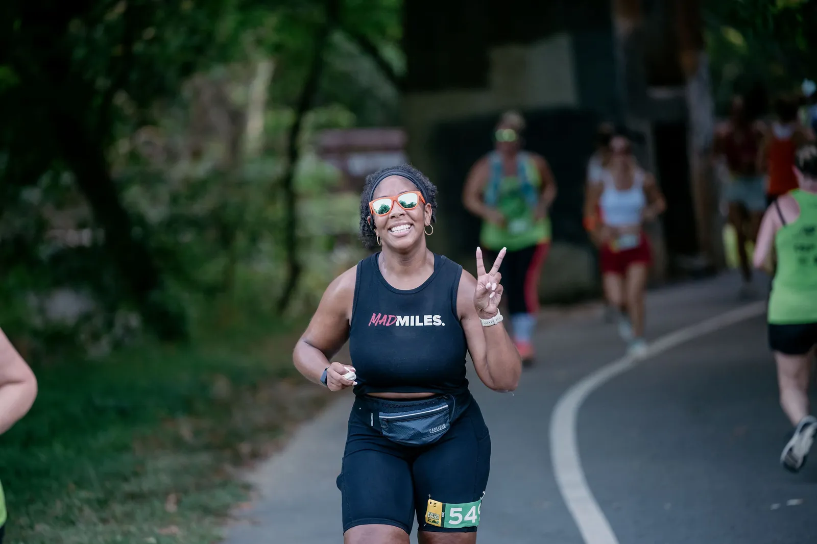 A smiling female runner wearing sunglasses and a black tank top with 'MADMILES' logo, gives a peace sign while running on a paved path.