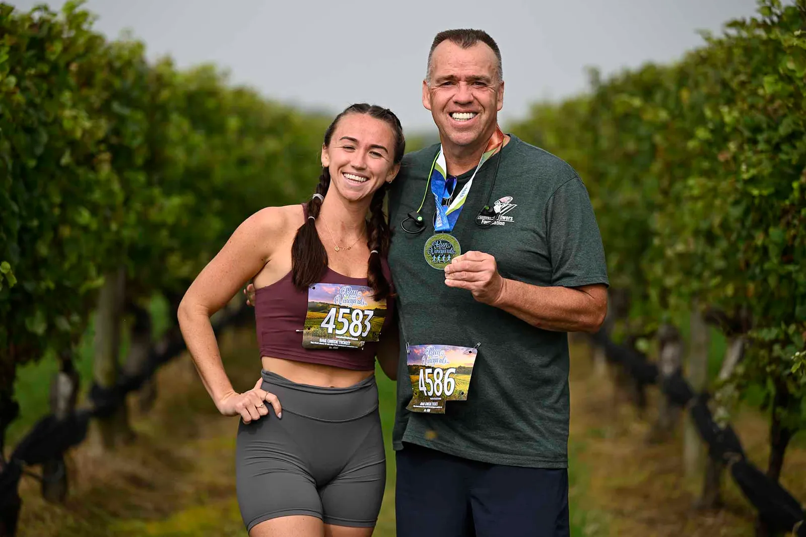 A man and a woman smile at the camera, both wearing race medals and bibs, with a vineyard in the background.