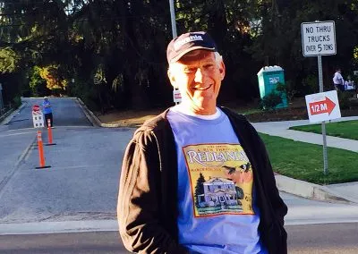 A smiling man wearing a 'Run Through Redlands' t-shirt and a baseball cap stands on a road with race signage and cones in the background.