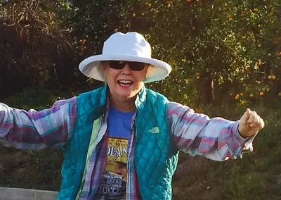 A smiling woman wearing a white hat and sunglasses, a teal vest over a plaid shirt, and a blue t-shirt, with her arms raised in celebration.