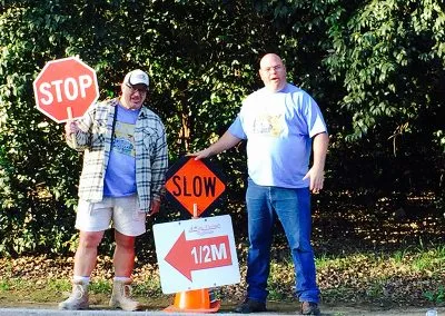 Two men stand by the side of the road, one holding a STOP sign and the other pointing to a sign indicating 1/2 mile and SLOW.