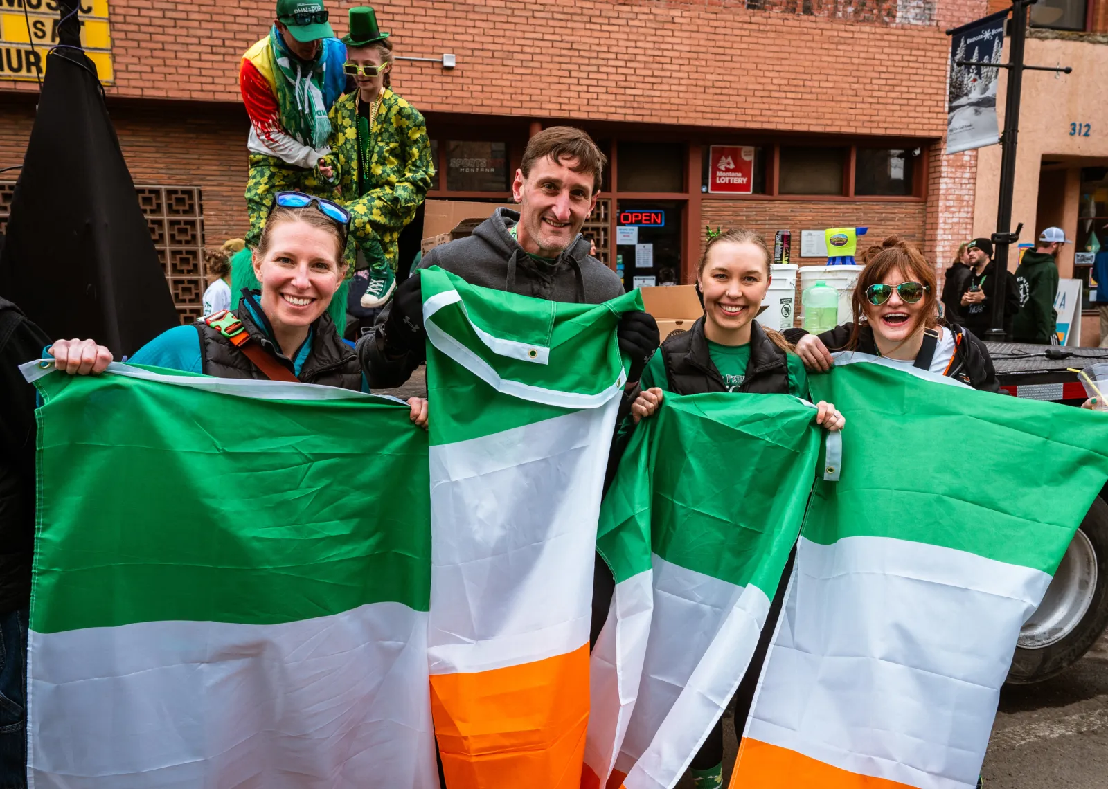 A group of smiling people holding Irish flags, with some dressed in festive green attire, suggesting a celebratory atmosphere at an event.