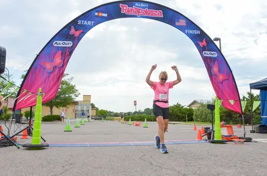 A female runner in a pink shirt and black shorts crosses the finish line with her arms raised in celebration under a large inflatable archway.