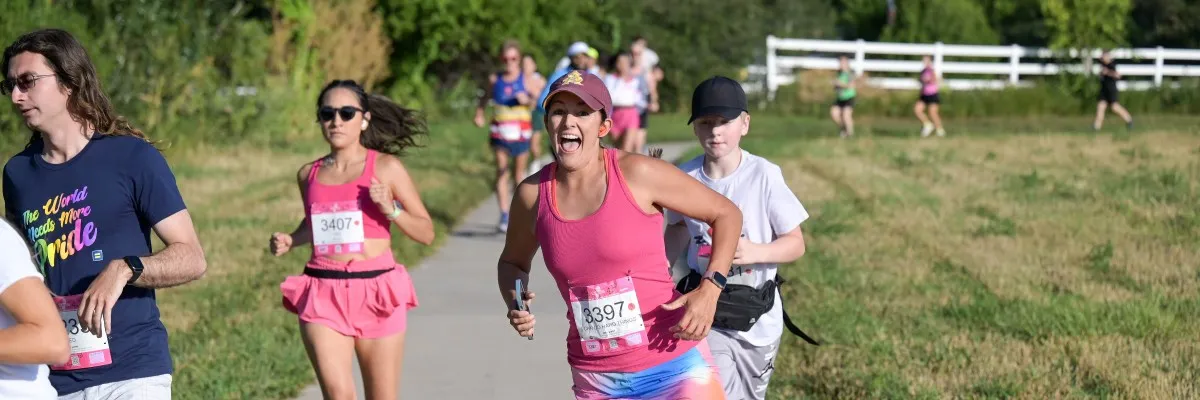 A group of runners, including a woman with an excited expression in the foreground, participate in a race on a paved path with grassy areas on either side.