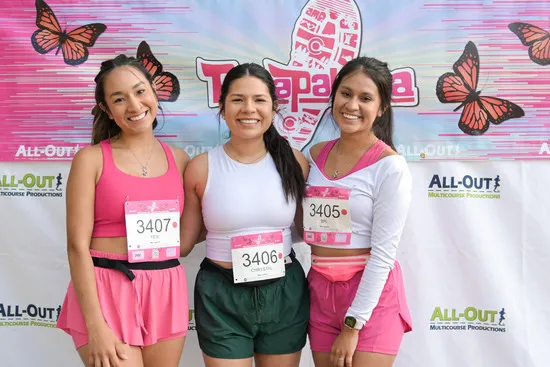 Three smiling women in running attire pose for a photo in front of a banner with butterflies and race branding.