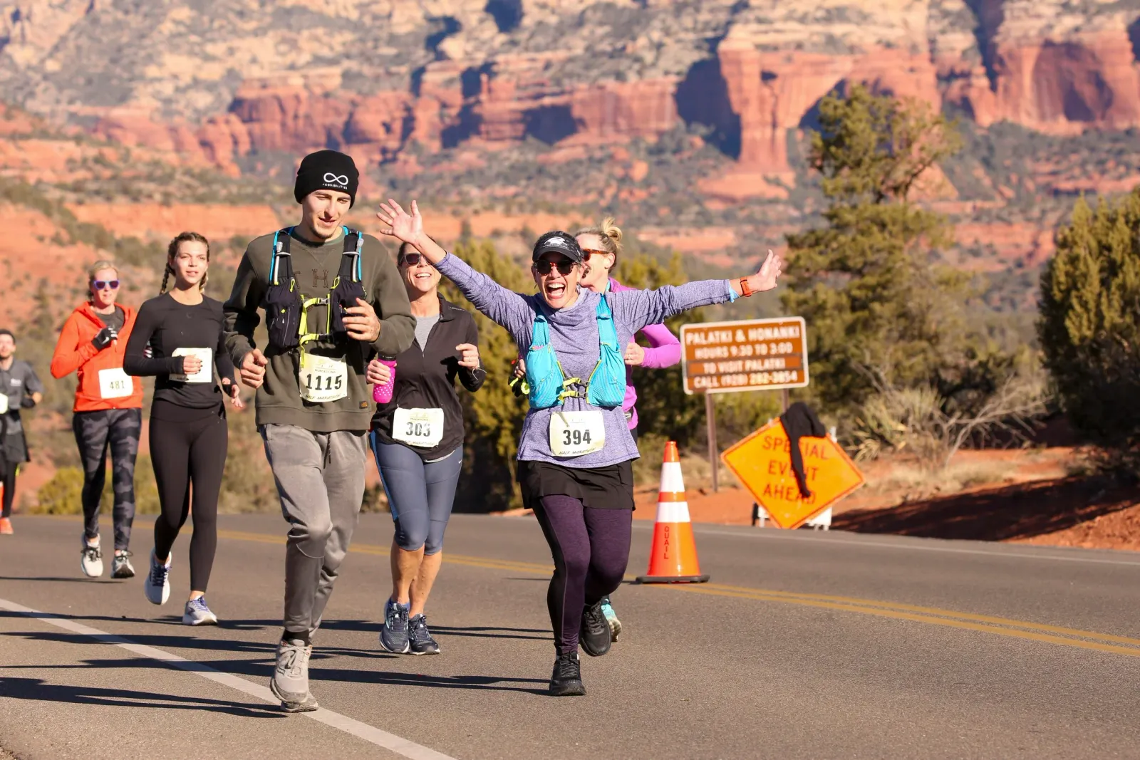 Runners participating in a half marathon with scenic red rock formations in the background. One runner in the foreground is smiling and waving with her arms outstretched.