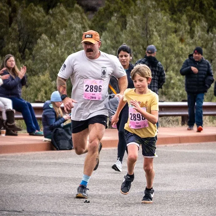 An adult runner and a child runner race side-by-side on a road, with spectators in the background.