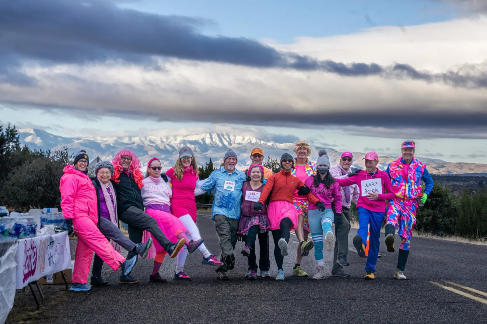 A group of runners in colorful costumes, including pink outfits and wigs, pose for a photo on a road with snowy mountains in the background.