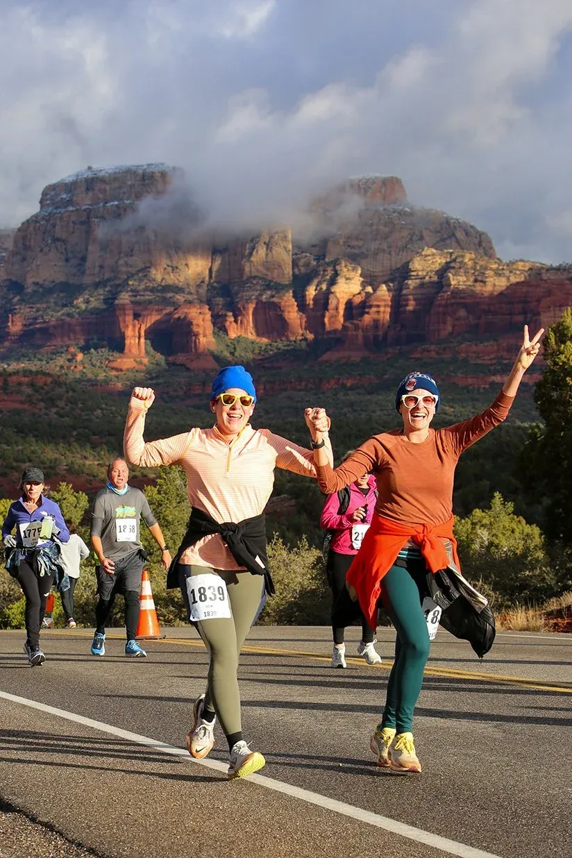 Two female runners celebrating with raised fists and a peace sign during a race, with dramatic red rock mountains in the background.